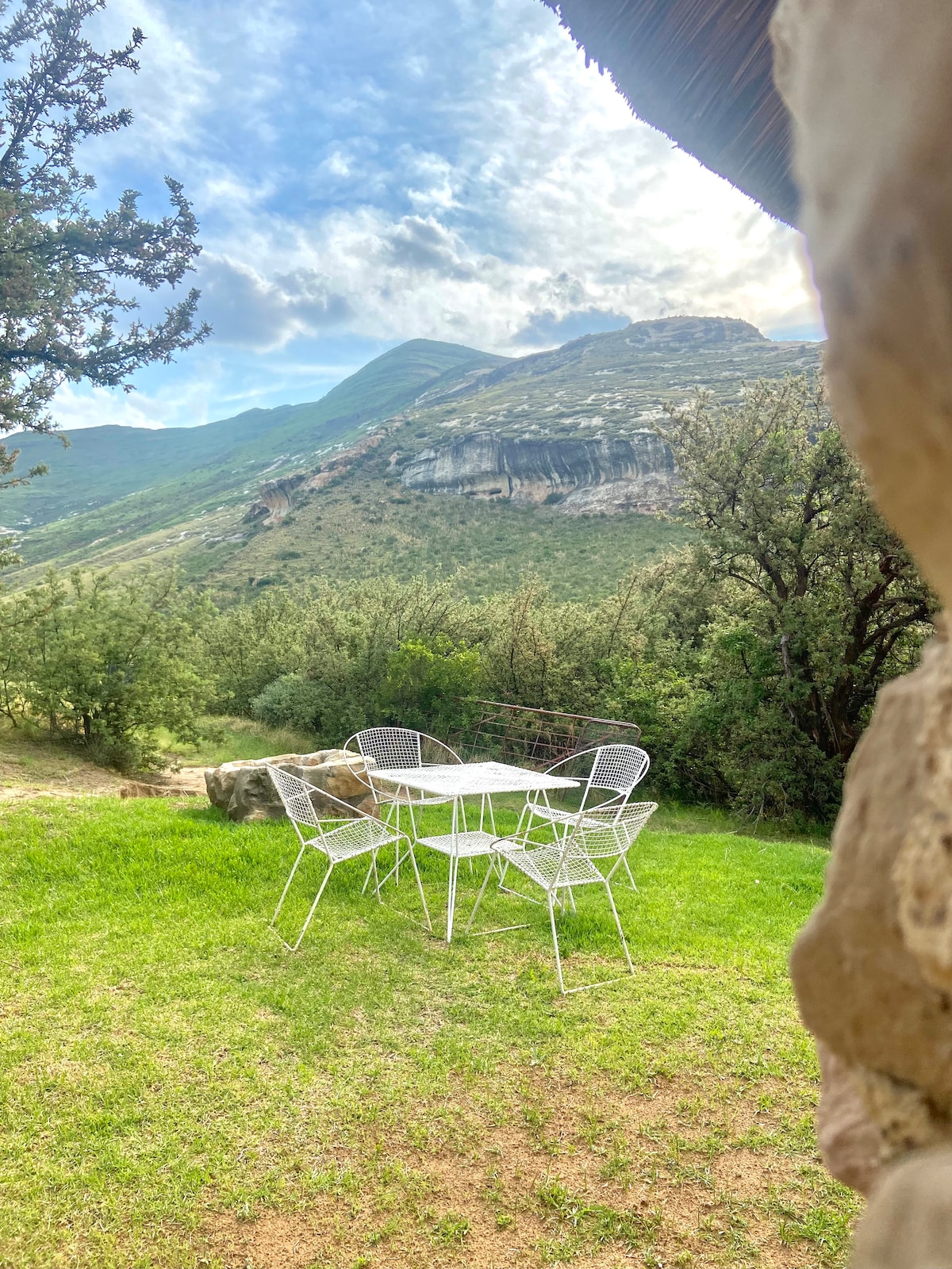 A quaint outdoor seating area is visible, featuring a white table surrounded by four chairs on a green lawn. The backdrop includes rolling hills and dramatic mountain scenery, with a partly cloudy sky overhead.
