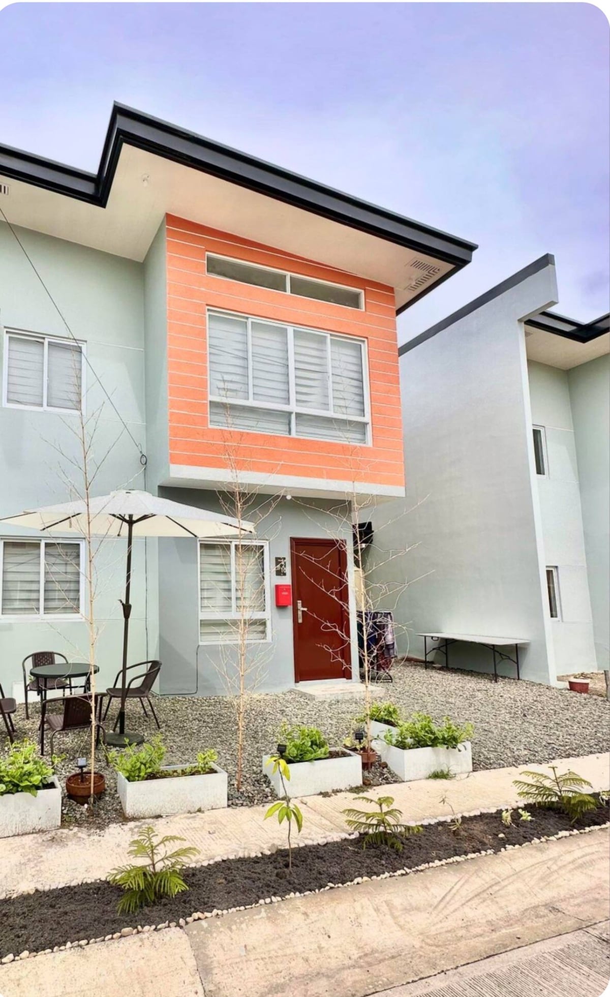 A two-story house is shown with a modern facade featuring a combination of light green and orange colors. A small outdoor area is visible with a white umbrella over a table and chairs, surrounded by minimalist landscaping and decorative plants.