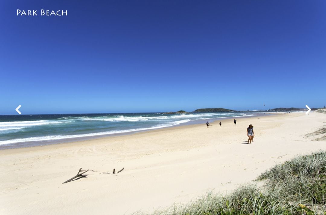A wide sandy beach extends under a clear blue sky, with gentle waves rolling onto the shore. People are seen walking along the water's edge, with grassy dunes in the foreground and distant hills along the coastline.