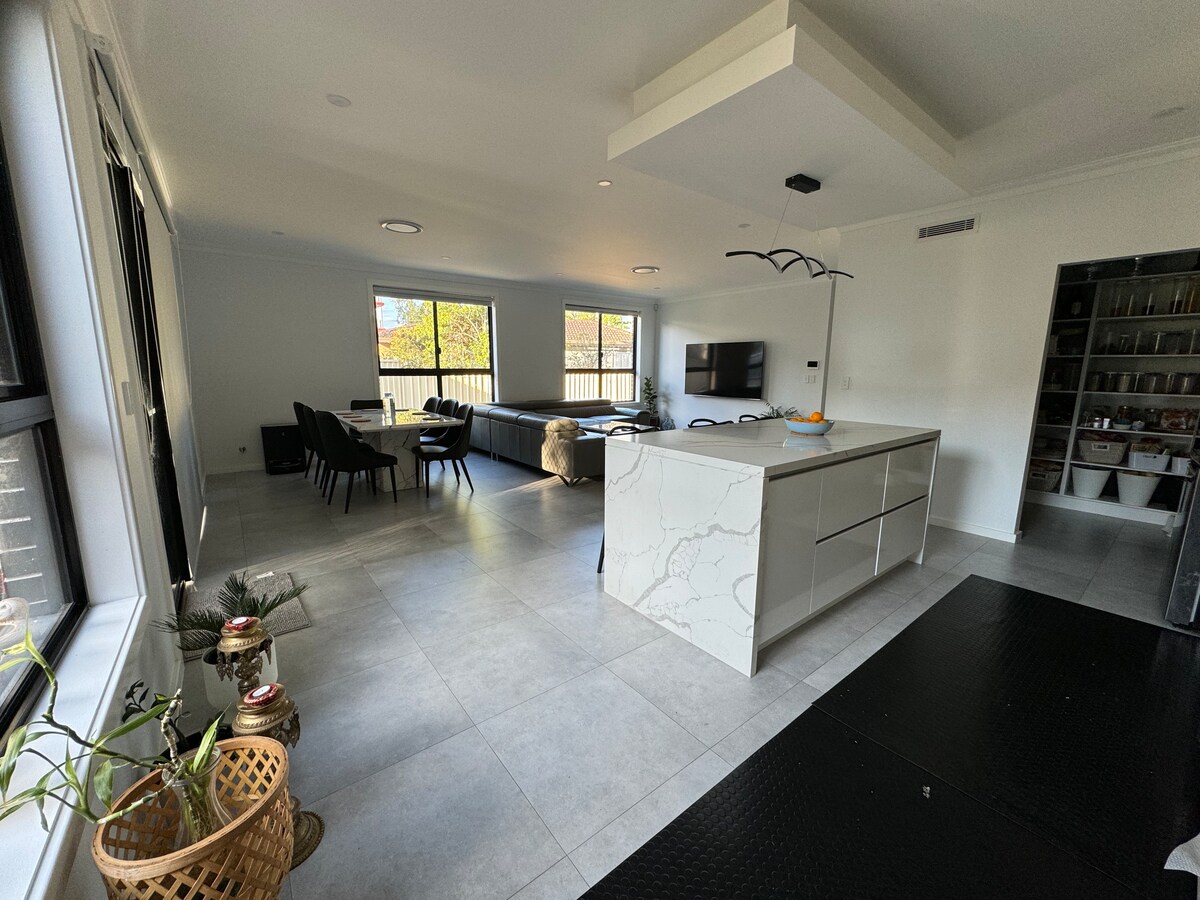 A spacious living area is highlighted by a central island with a marble-like surface. Large windows allow natural light to fill the room, which features a comfortable seating arrangement and a modern television mounted on the wall. Indoor plants add a touch of greenery.