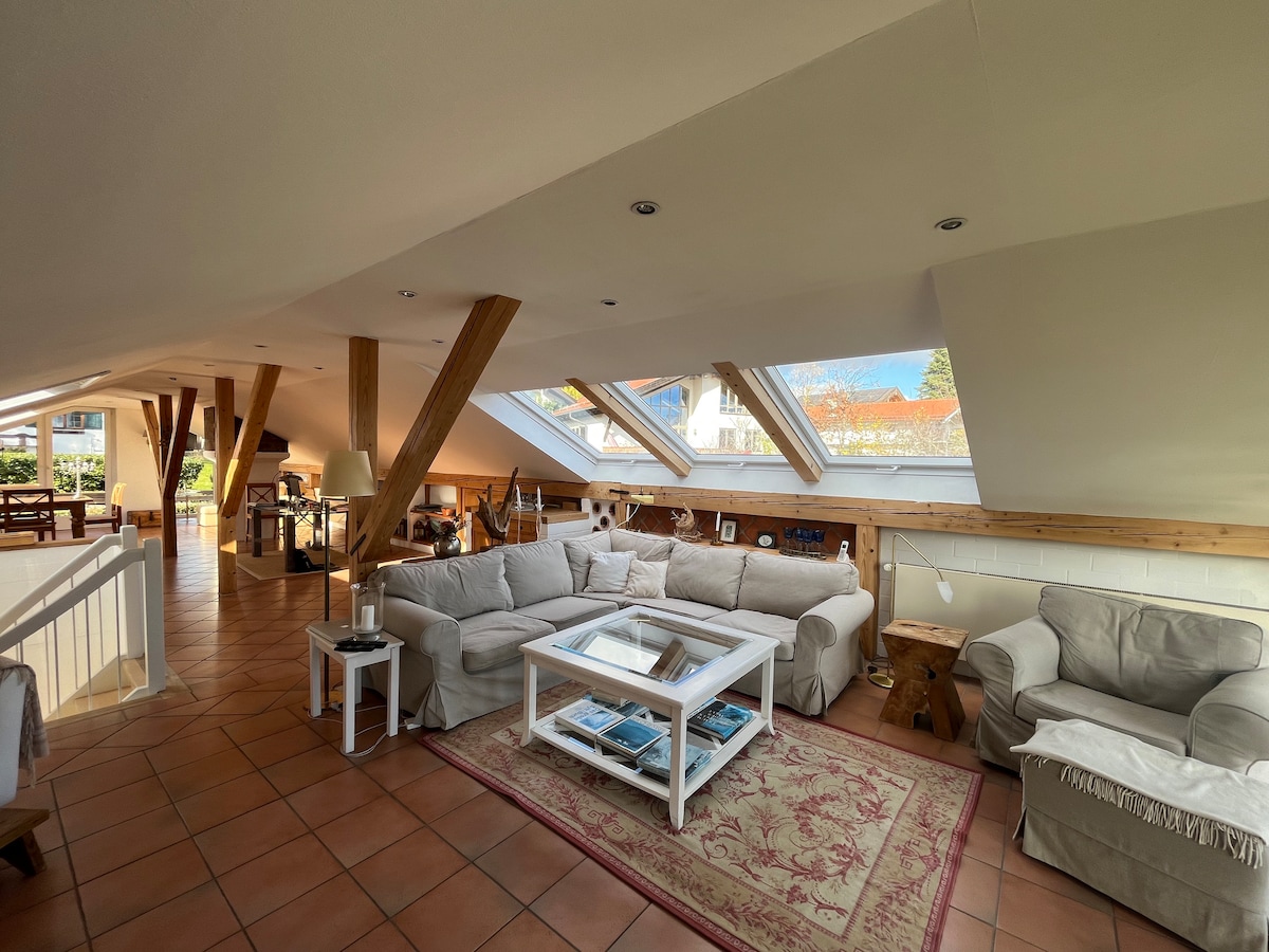 A spacious living area is highlighted by slanted skylights that allow natural light to fill the room. Comfortable seating options surround a central coffee table, while a subtle rug adds warmth to the tiled floor. Wooden beams accent the ceiling, enhancing the open layout.