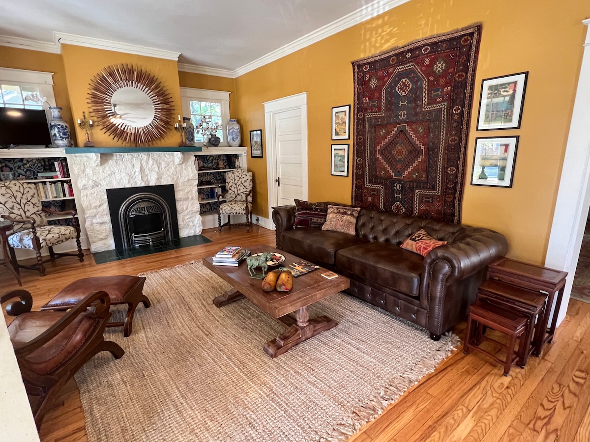 A warm living room features a leather sofa facing a stone fireplace, with bookshelves and decorative items on either side. A woven rug lays beneath a wooden coffee table, which is adorned with fruit and books. Natural light brightens the space through nearby windows.