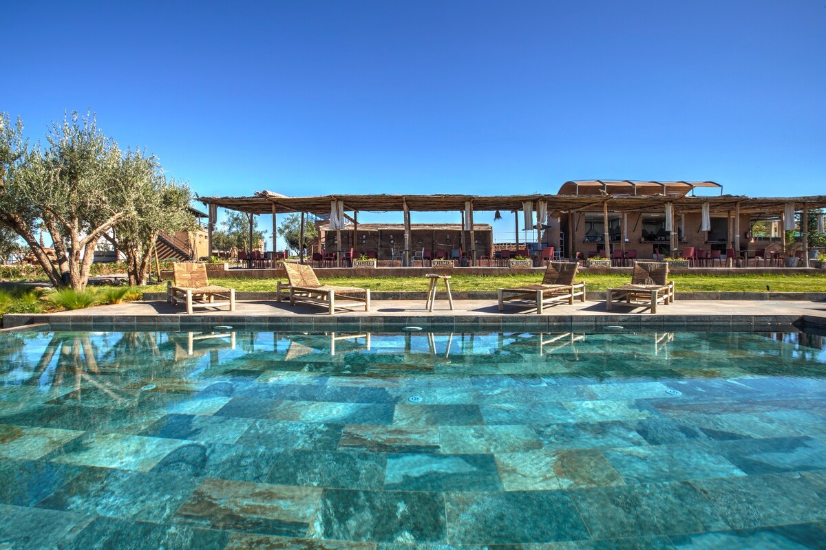 A serene pool area is framed by lush greenery, featuring a clear blue water surface that reflects the bright sky. Comfortable lounge chairs are positioned around the pool, while a spacious outdoor dining area with shaded seating is visible in the background.