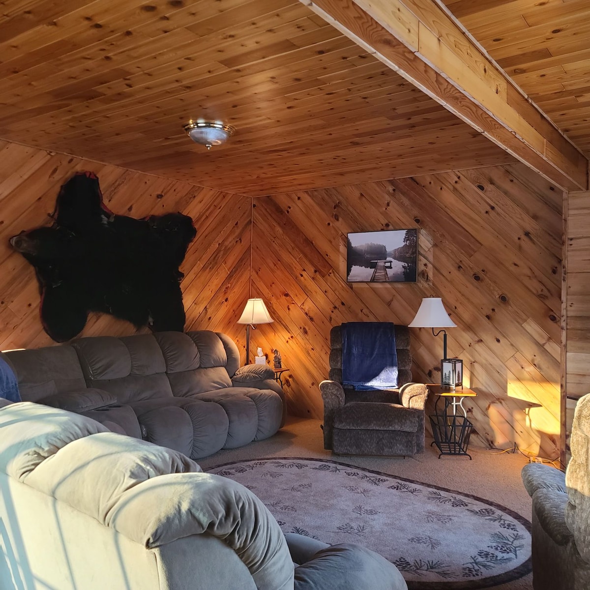 A cozy seating area features a large sectional sofa and a plush armchair with a side table. Natural wood paneling complements the soft lighting from table lamps. A decorative rug lies on the carpeted floor, enhancing the inviting ambiance of the space.