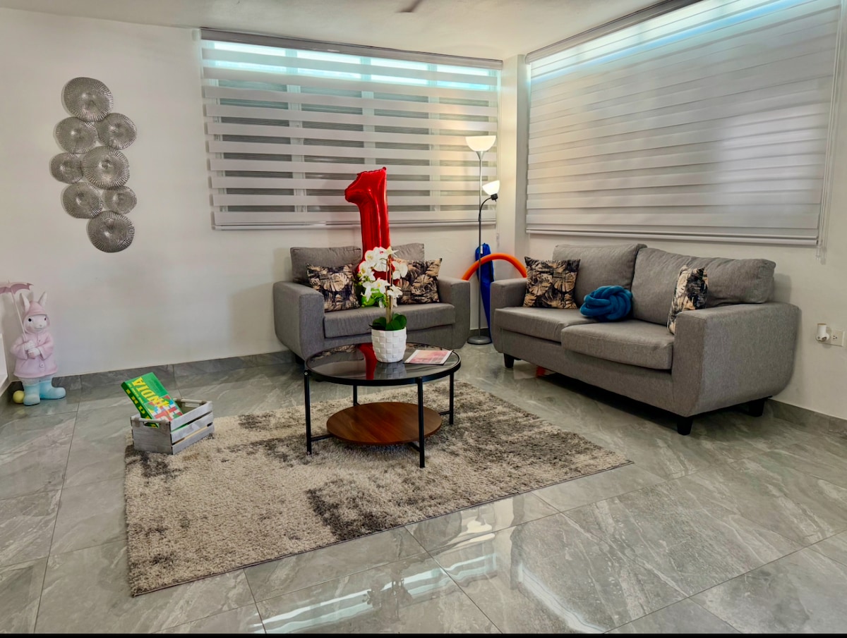 A bright living area features a gray sofa paired with a cushioned sectional. A coffee table sits atop a soft rug, surrounded by decorative items. Natural light filters through modern window shades, complementing the serene color palette. An additional lamp provides ambient lighting.