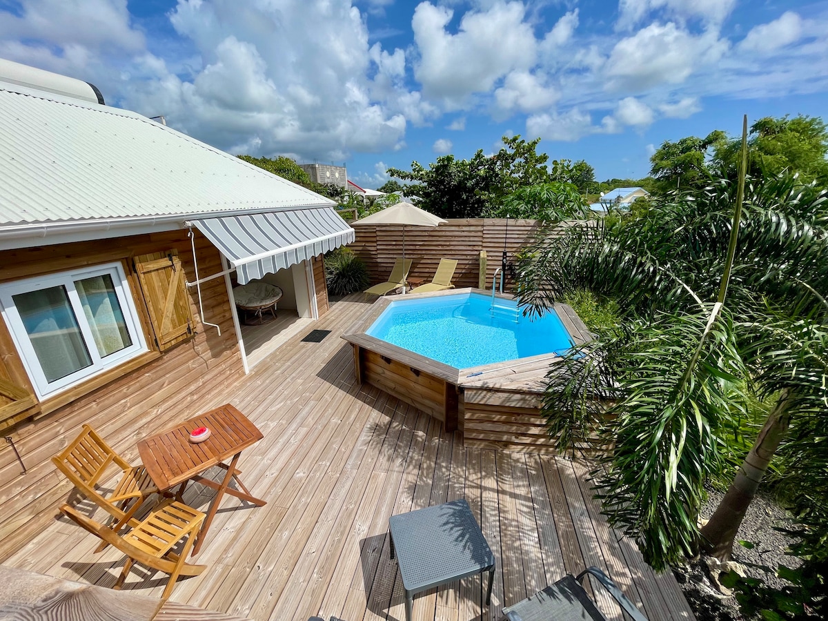 A private pool is situated on a wooden deck, surrounded by tropical foliage. Two lounge chairs rest beside the pool, and a small dining table with two chairs is placed nearby. The bungalow features a light-colored façade under a partly cloudy sky.