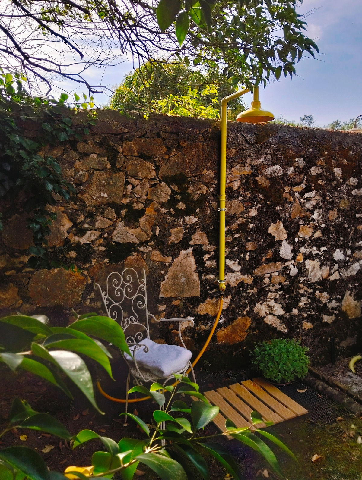 An outdoor shower with a yellow fixture is mounted against a textured stone wall, surrounded by lush greenery. A white garden chair is positioned nearby on a wooden mat, creating a relaxing outdoor space.