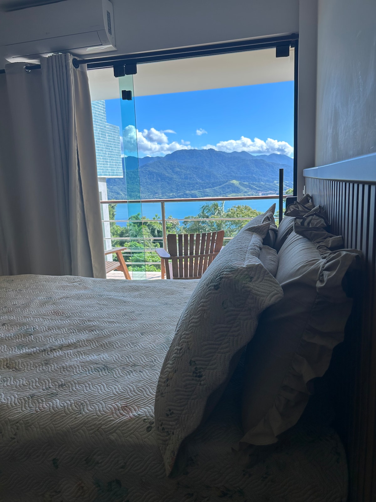 A tranquil sleeping area is shown with a spacious bed featuring decorative pillows. A large window offers a view of verdant mountains and a shimmering body of water, while a wooden balcony set is visible outside, inviting relaxation.