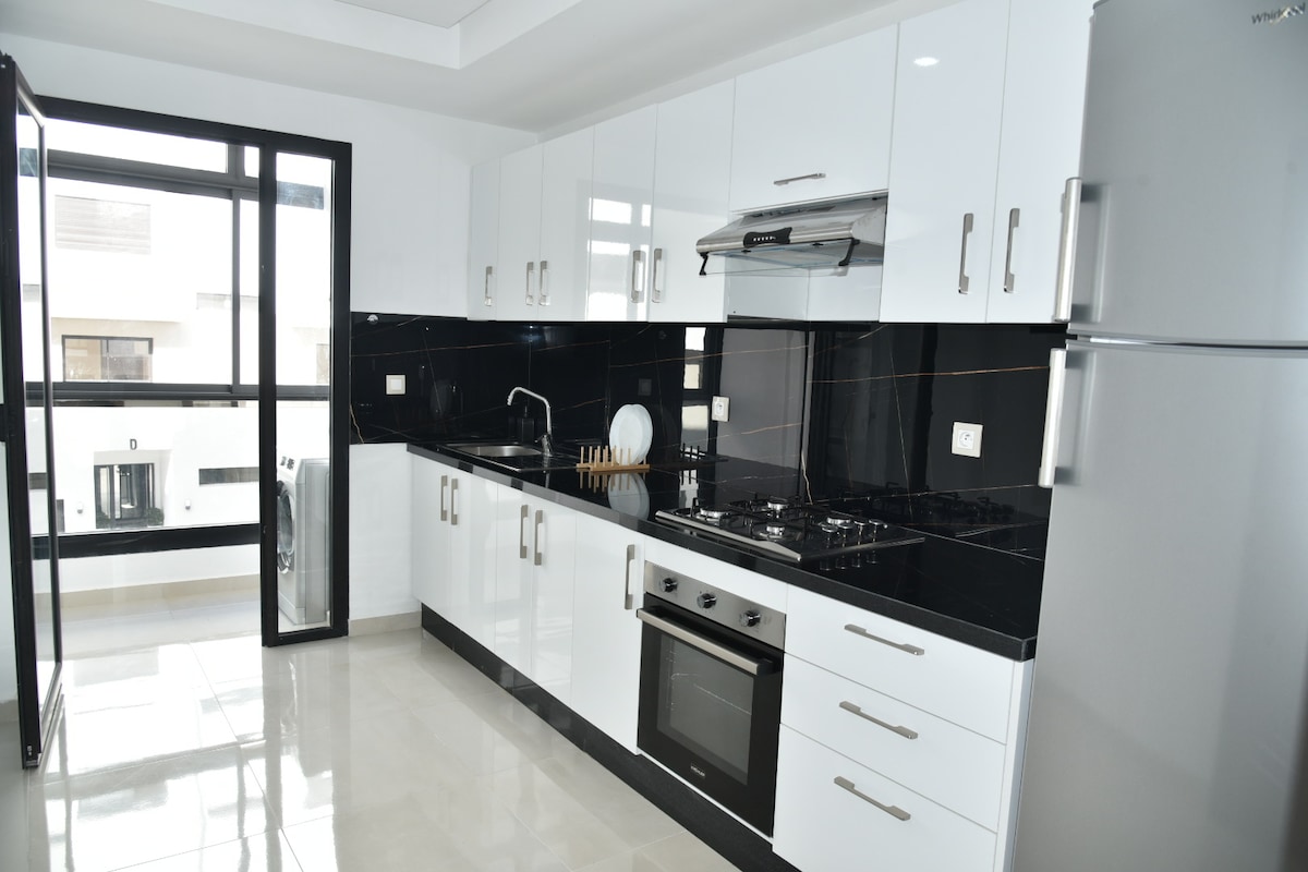 A modern kitchen features sleek white cabinetry paired with dark countertops. Stainless steel appliances, including a refrigerator and oven, are integrated into the space. Large windows allow natural light to illuminate the area, enhancing the functionality and aesthetics of the kitchen.