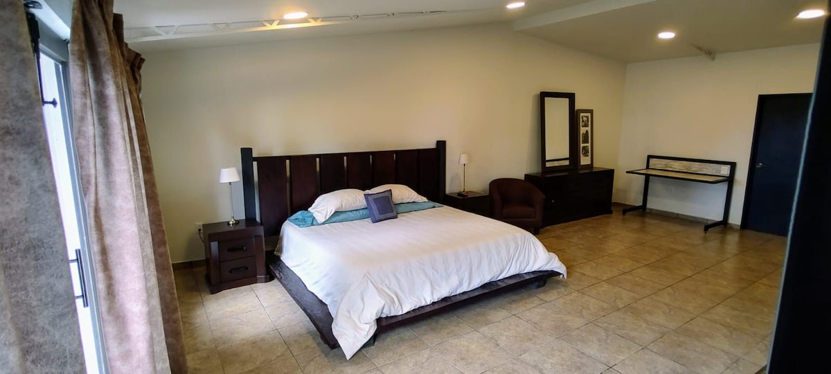 A spacious sleeping area features a large bed with a light blanket and an accent pillow. Dark wood furniture, including a nightstand and dresser, is present. Natural light enters through a window with beige curtains, complementing the tiled floor and neutral wall color.