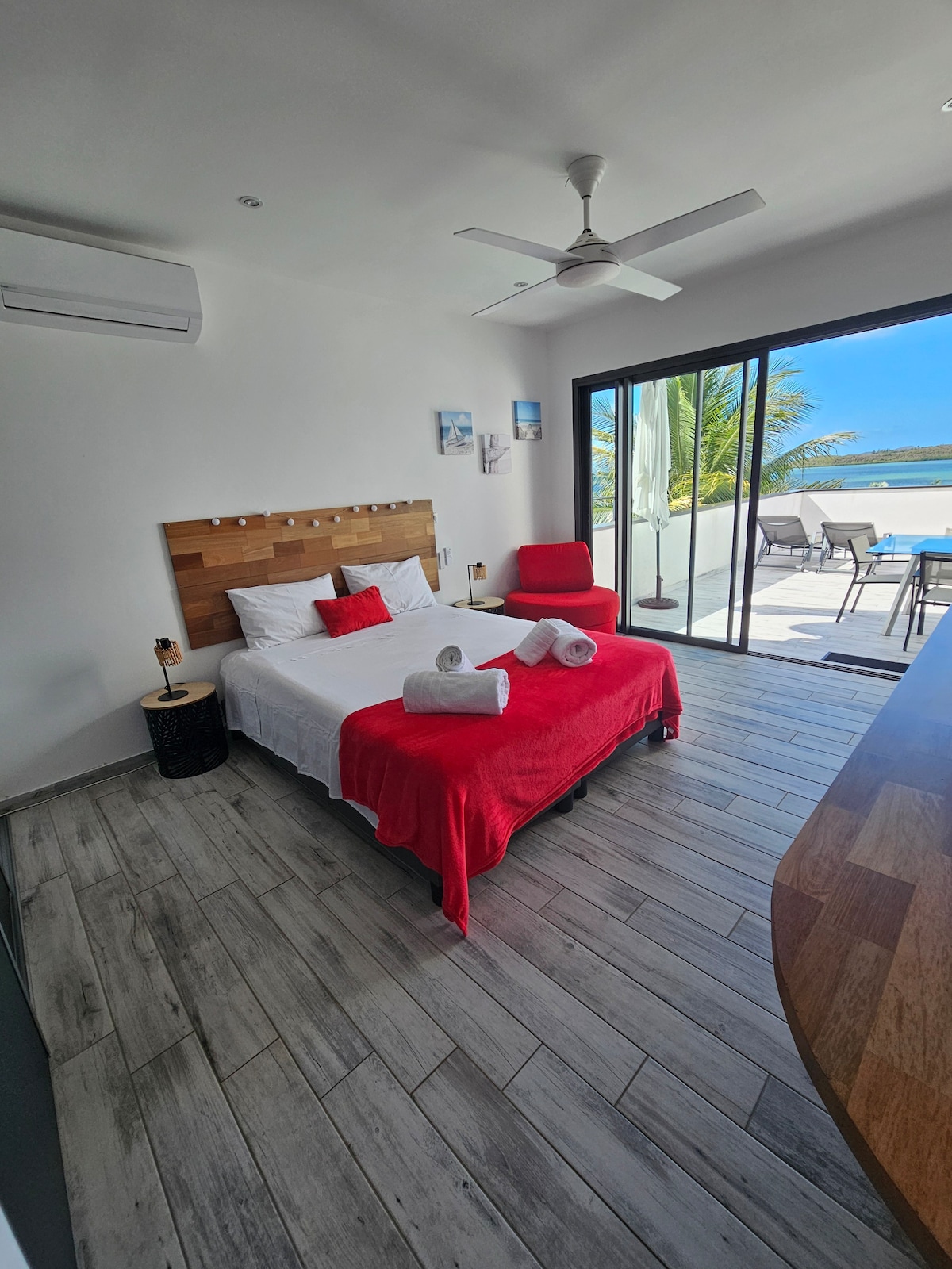 A bright, airy bedroom features a spacious bed dressed in white linens, complemented by a vibrant red throw. Large glass doors open to a patio area, revealing views of the outdoor seating and serene surroundings. Decorative elements include red accents and a ceiling fan.