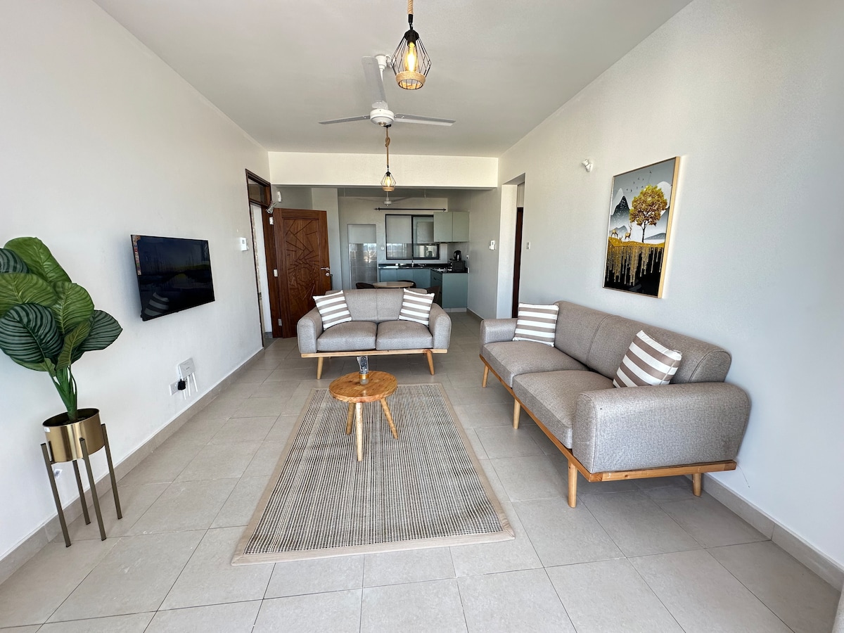 A bright living area features two modern sofas with striped cushions, positioned around a round wood coffee table. A textured area rug is placed on the tiled floor. A ceiling fan and wall-mounted TV are included, with open views toward the kitchen area.