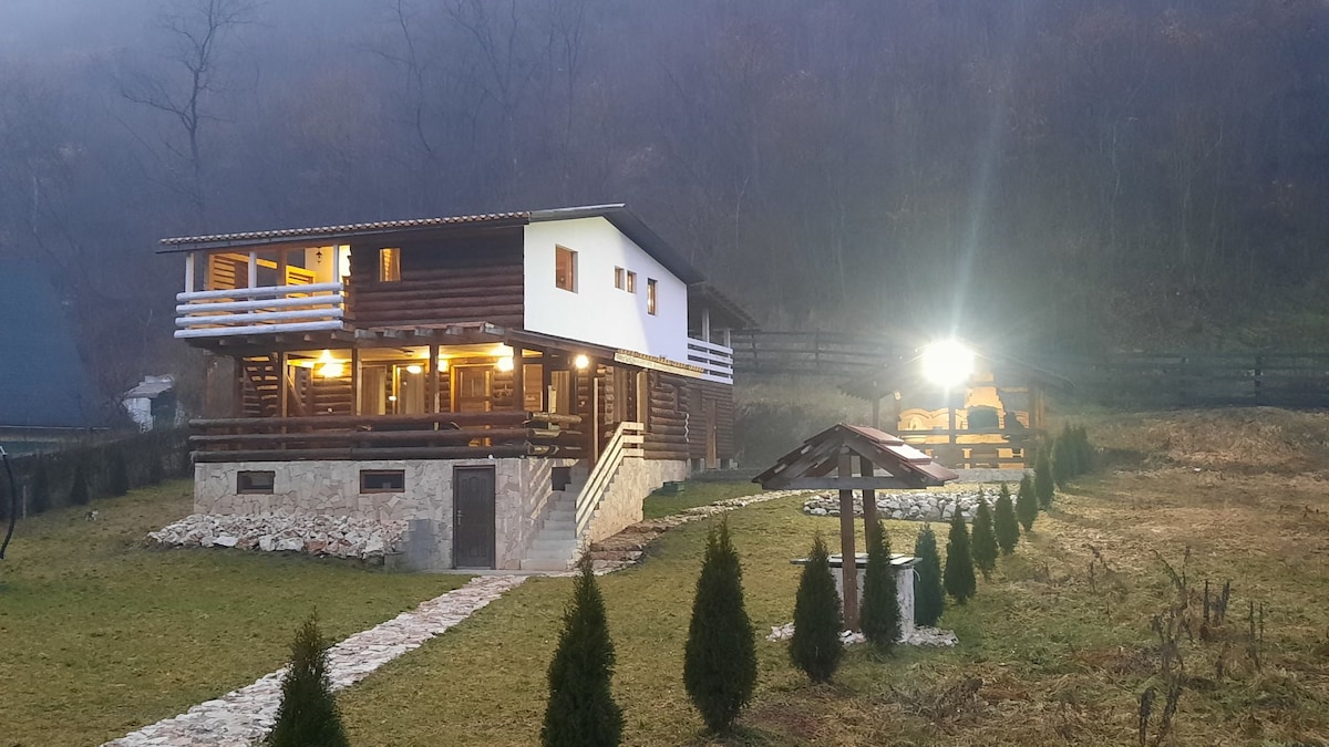The exterior of a spacious cabin is shown, featuring a mix of wooden and white elements. A well-maintained lawn with neatly trimmed shrubs leads to a stone pathway. Soft lighting is visible, creating a welcoming atmosphere, and the surrounding landscape is partially shrouded in mist.