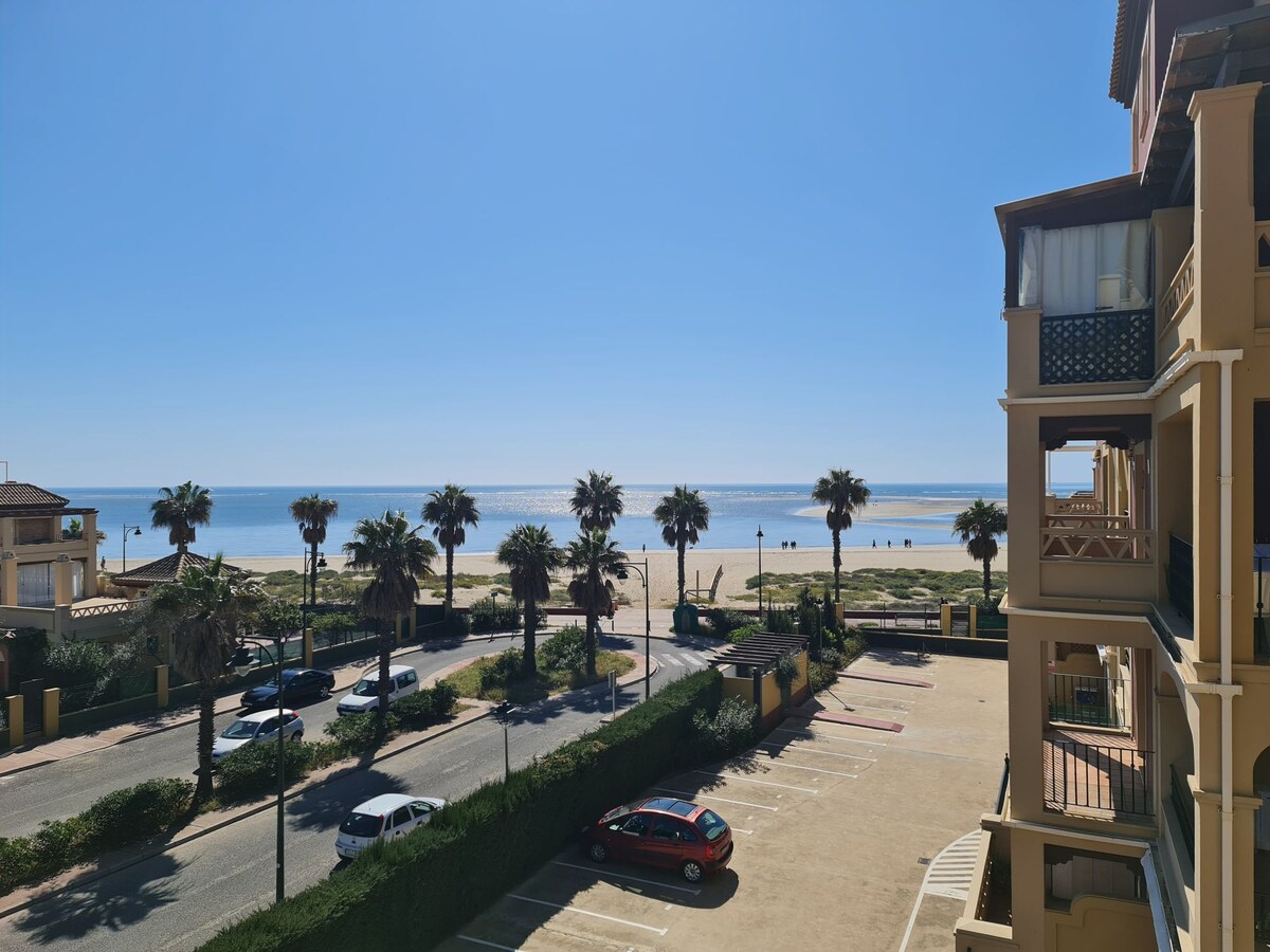 A serene view from the balcony reveals a sandy beach lined with palm trees, glistening under bright sunshine. A calm sea stretches towards the horizon, while a parking area with colorful cars is visible below, enhancing the coastal setting.