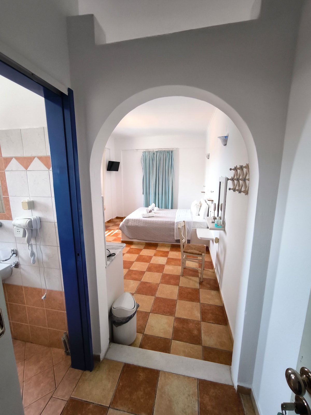 An inviting entrance leads into a spacious room showcasing a light-colored bed and soft drapes. The area features a small table and chair, with tiled floors providing a warm touch. Visible are glimpses of a nearby bathroom and well-lit space, enhancing the overall tranquility.