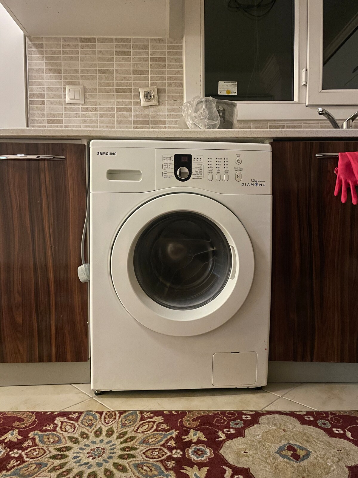 A white washing machine is positioned against a wooden cabinet in a well-lit kitchen. The machine features a round door with a transparent window, revealing a drum inside. A colorful area rug with intricate patterns covers the floor beneath.
