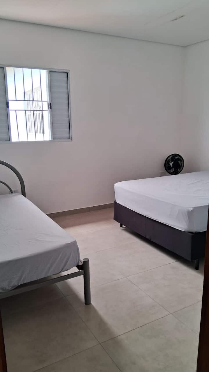 Druhá spálňa domu na druhom poschodí s manželskou posteľou, samostatnou posteľou, vežovým ventilátorom a stojanom na batožinu.