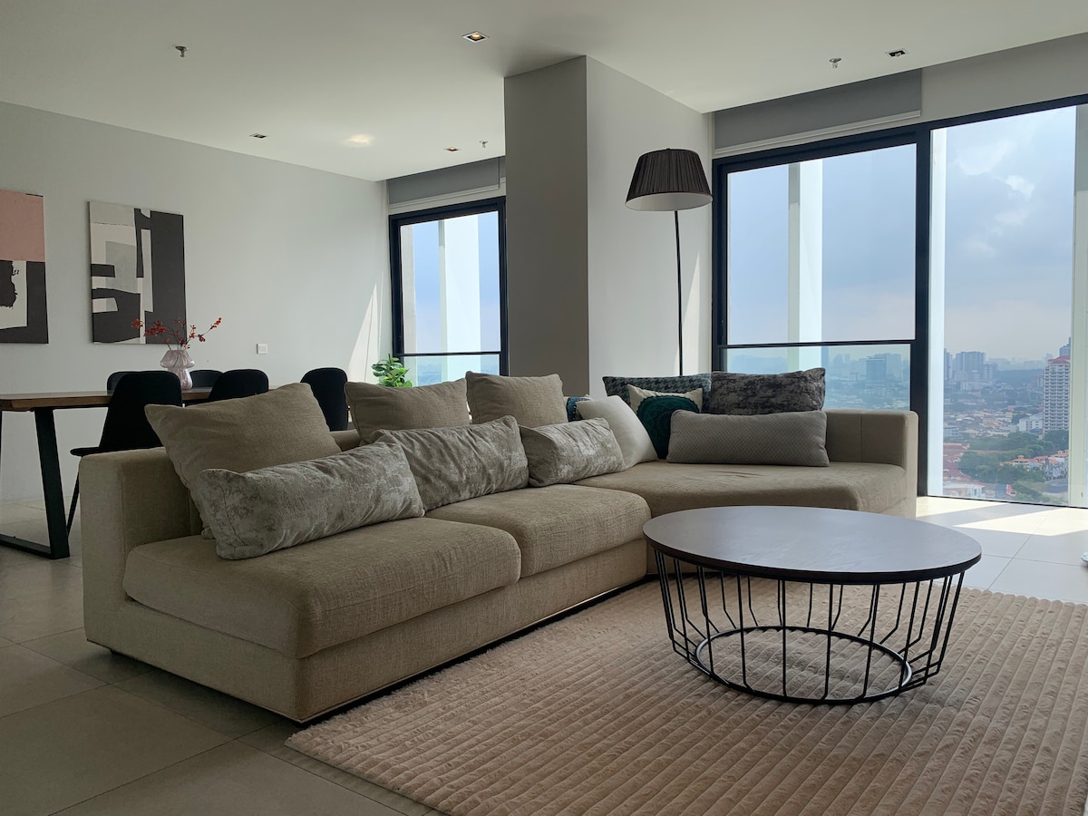 A spacious living area features a large, L-shaped sofa adorned with decorative cushions. A round coffee table with a wireframe base sits on a soft area rug. Floor-to-ceiling windows provide scenic views of the surrounding city and ample natural light.