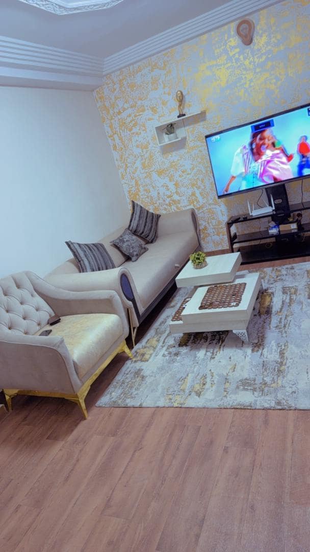 A cozy living area features a light-colored, tufted sofa alongside a matching armchair. A low coffee table sits on a patterned rug, while a television is mounted on the wall, providing entertainment options. Warm lighting enhances the relaxed ambiance.