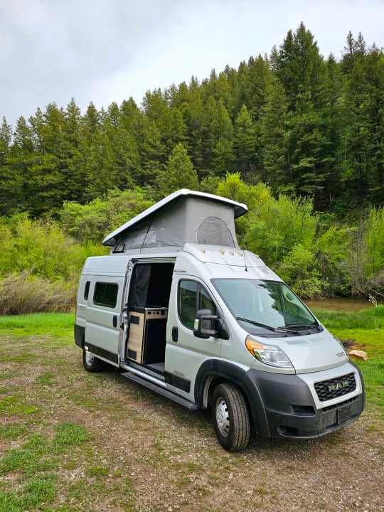 A compact motorhome is parked in a grassy area surrounded by lush green trees. The vehicle features a pop-top roof for additional space and a sliding side door that opens to reveal a stylish interior. Natural light enhances the exterior's modern design.