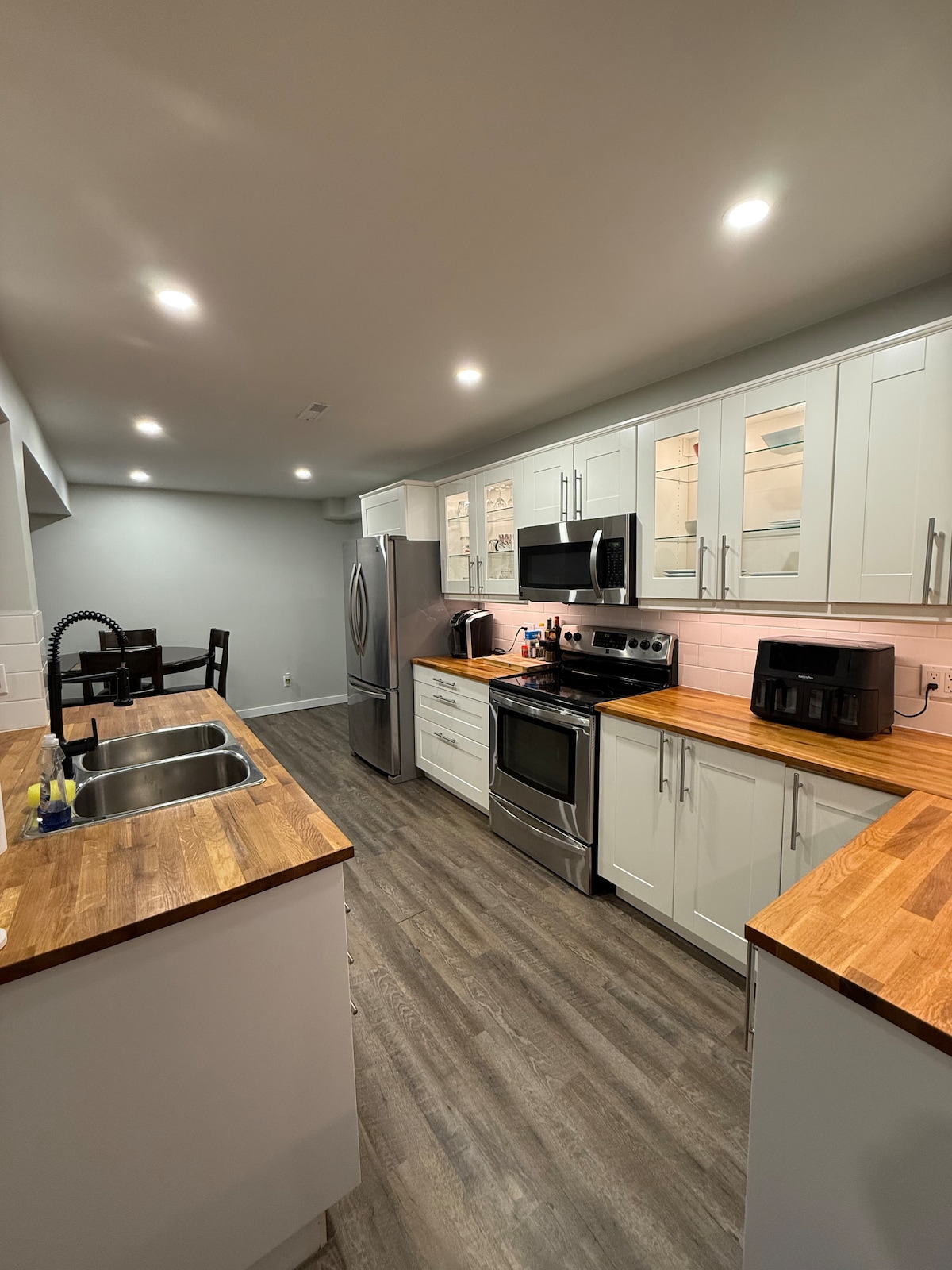 A spacious kitchen features modern cabinetry with glass accents and wooden countertops. Stainless steel appliances, including a refrigerator and stove, are prominently displayed. Under-cabinet lighting illuminates the workspace, while a small dining area is visible in the background.