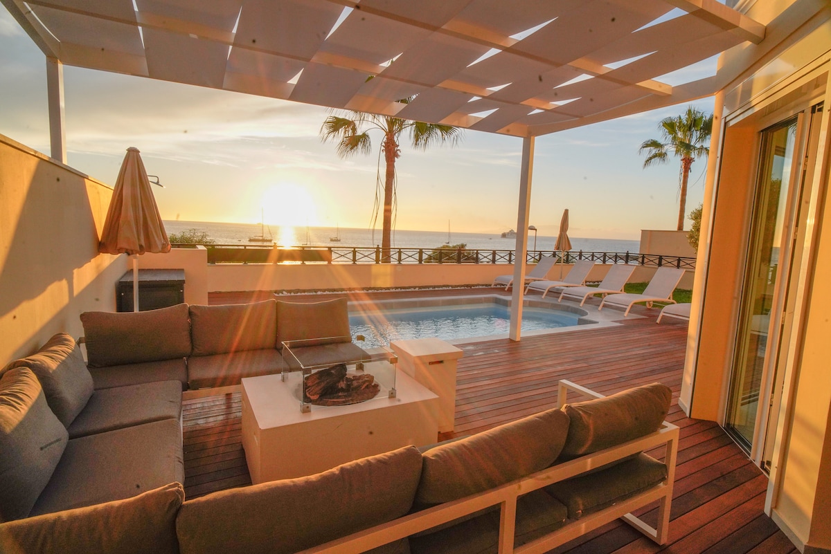 An outdoor lounge area features a large sectional sofa around a fire pit, with the sun setting over the sea in the background. The space is framed by palm trees, and a shimmering pool reflects the golden light.