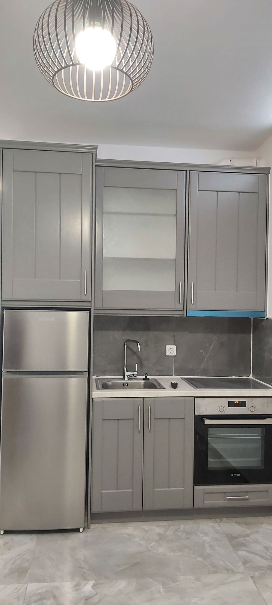 A modern kitchen is depicted, showcasing grey cabinetry with glass panel accents. Stainless steel appliances, including a refrigerator, oven, and a sink, are neatly integrated. The countertop features a minimalist design, complemented by neutral tiles on the wall and floor.