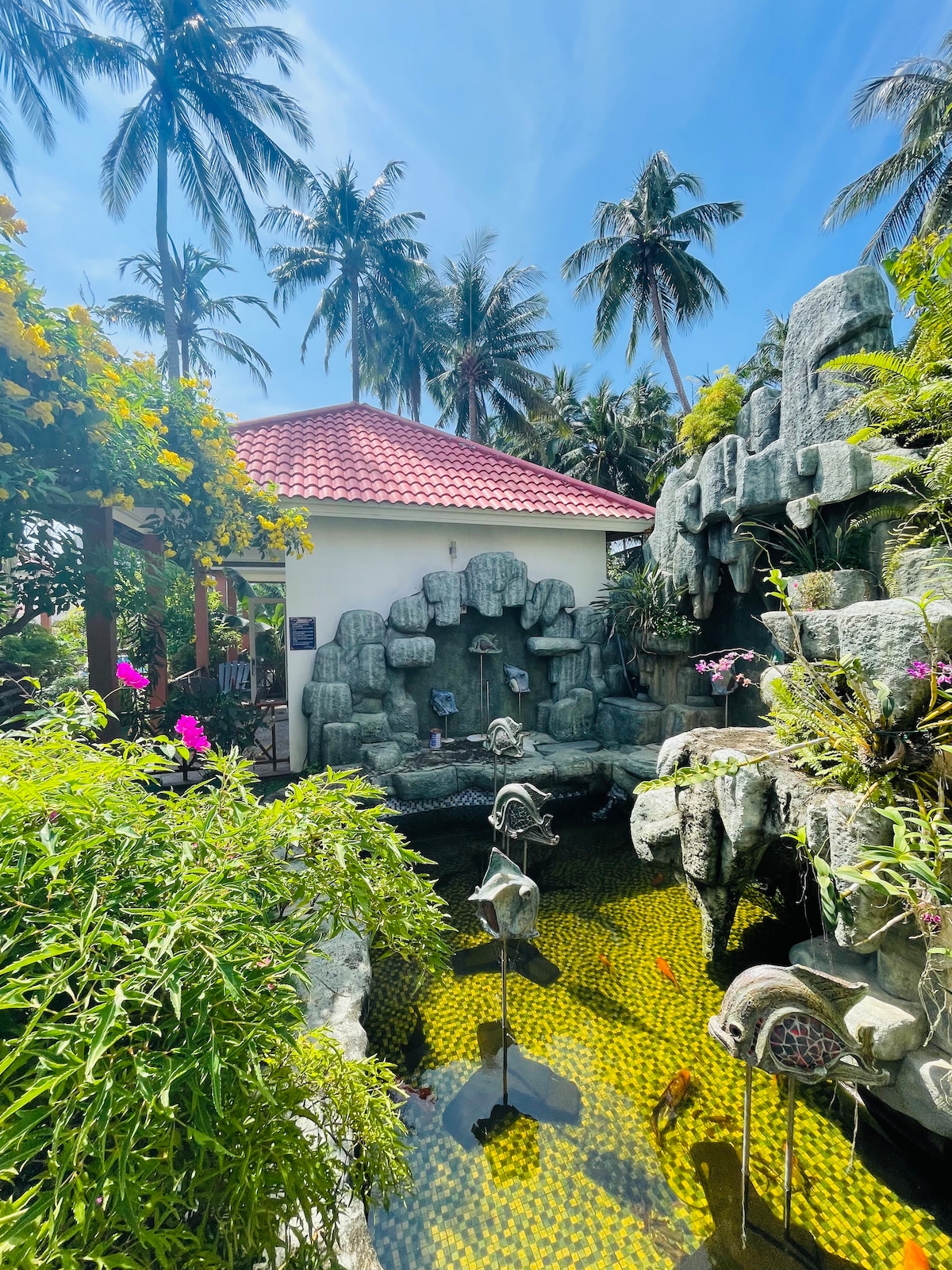A serene garden area features a sculpted rock pond adorned with decorative fish sculptures. Lush greenery and colorful flowers surround the space, while palm trees sway gently in the background. A charming bungalow with a red-tiled roof is visible, adding to the tranquil atmosphere.