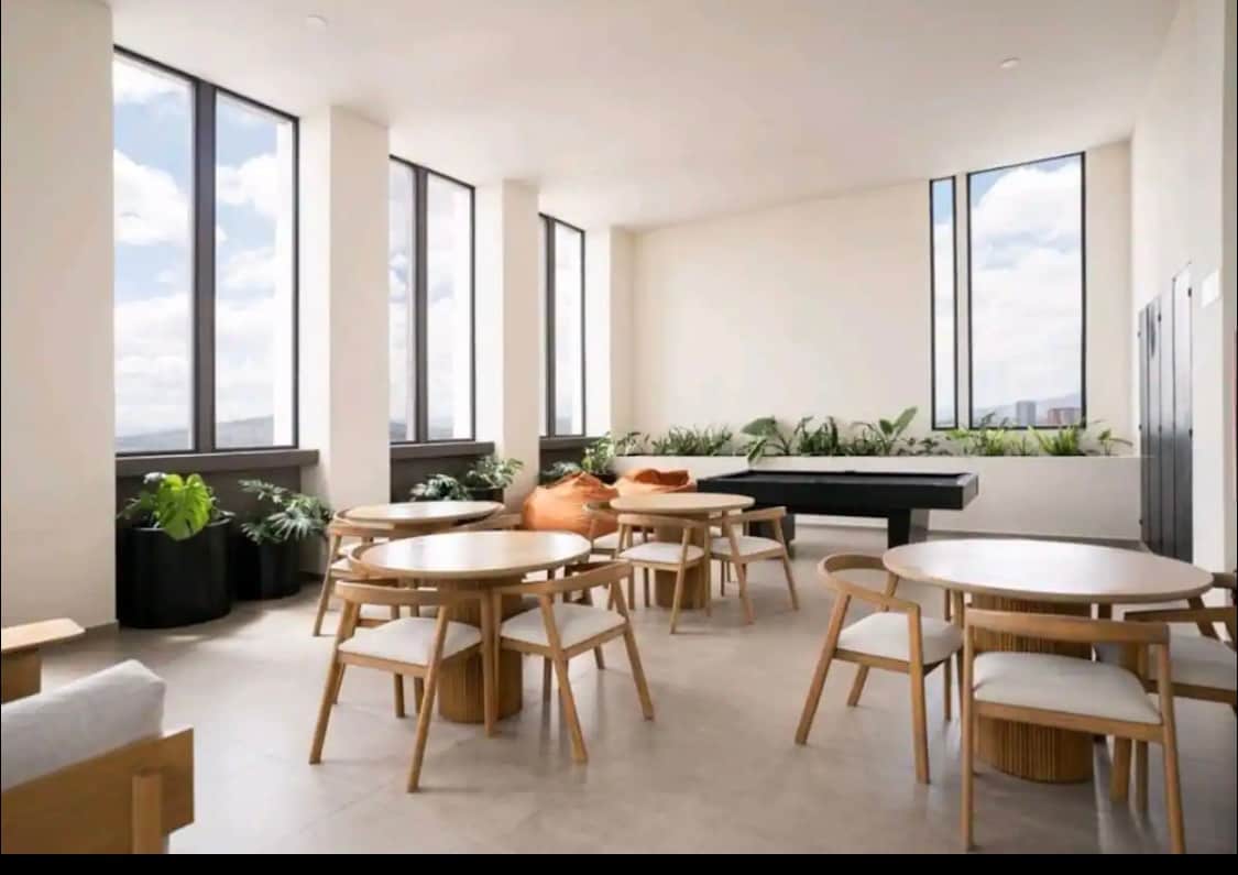 A shared common area features natural light streaming through large windows, illuminating several wooden tables with chairs. Lush green plants are positioned in black pots, adding a touch of nature to the space. A comfortable seating area is visible, inviting conversation.