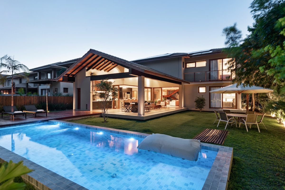A modern house is depicted with a spacious outdoor area featuring a rectangular pool surrounded by a manicured lawn. The open sliding doors reveal an interior space with dining and living areas. A shaded patio with seating is visible next to the pool, enhancing outdoor relaxation.