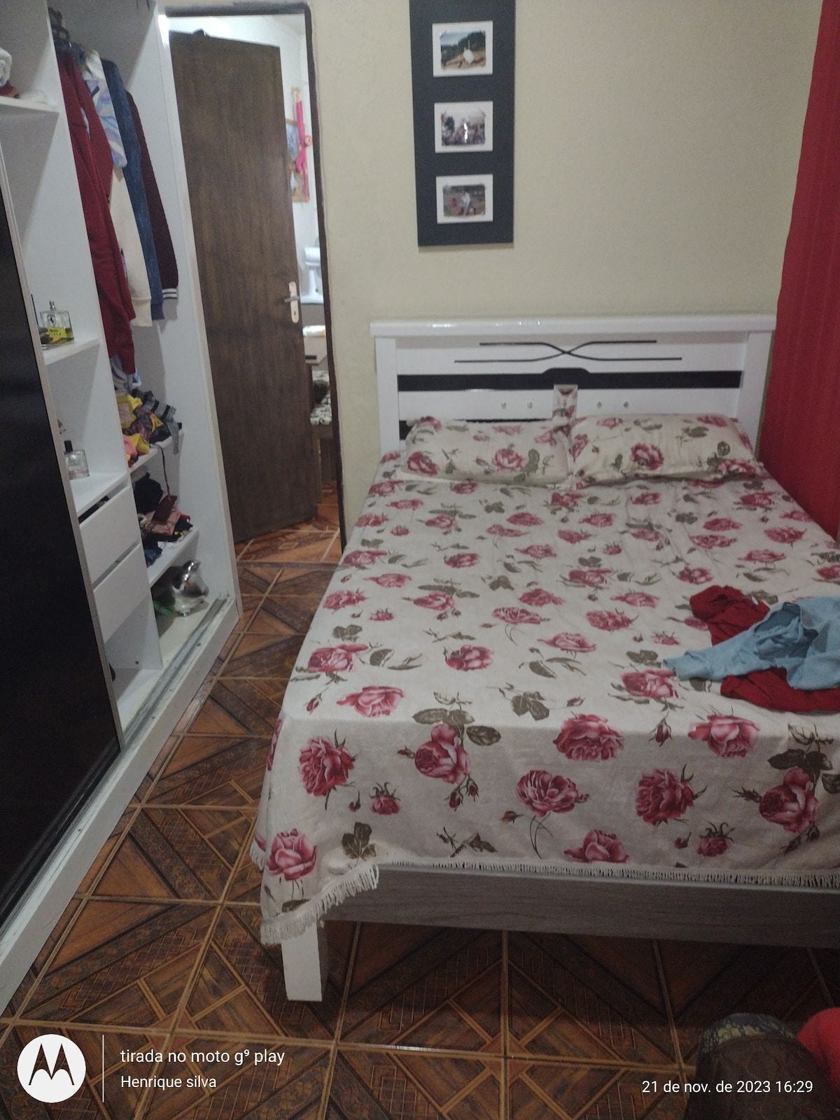 A cozy sleeping area is presented, featuring a floral-patterned bedspread covering a spacious bed. A wardrobe is visible along one wall, offering storage space. The entrance to the room is framed by a wooden door, while framed photos adorn the wall, adding a personal touch.