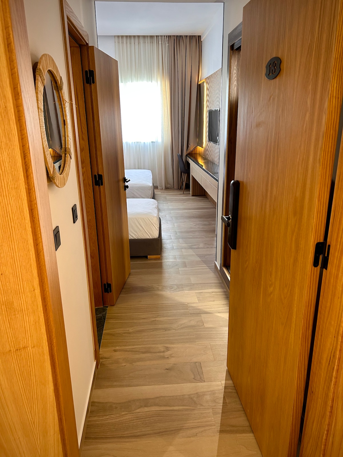 A hallway is depicted, leading to a room with two beds. Light wood doors frame the entrance, and a mirror is mounted on the wall. Natural light filters in through a window at the far end, illuminating the warm wooden flooring.