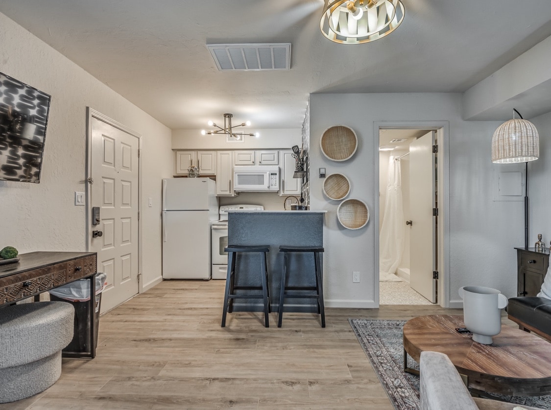 A well-appointed studio kitchen is equipped with essential appliances, including a refrigerator and stove. A dining area with two bar stools is positioned near the kitchen island. The light-colored walls and wood flooring create an open and inviting space, with a glimpse of the bathroom visible in the background.