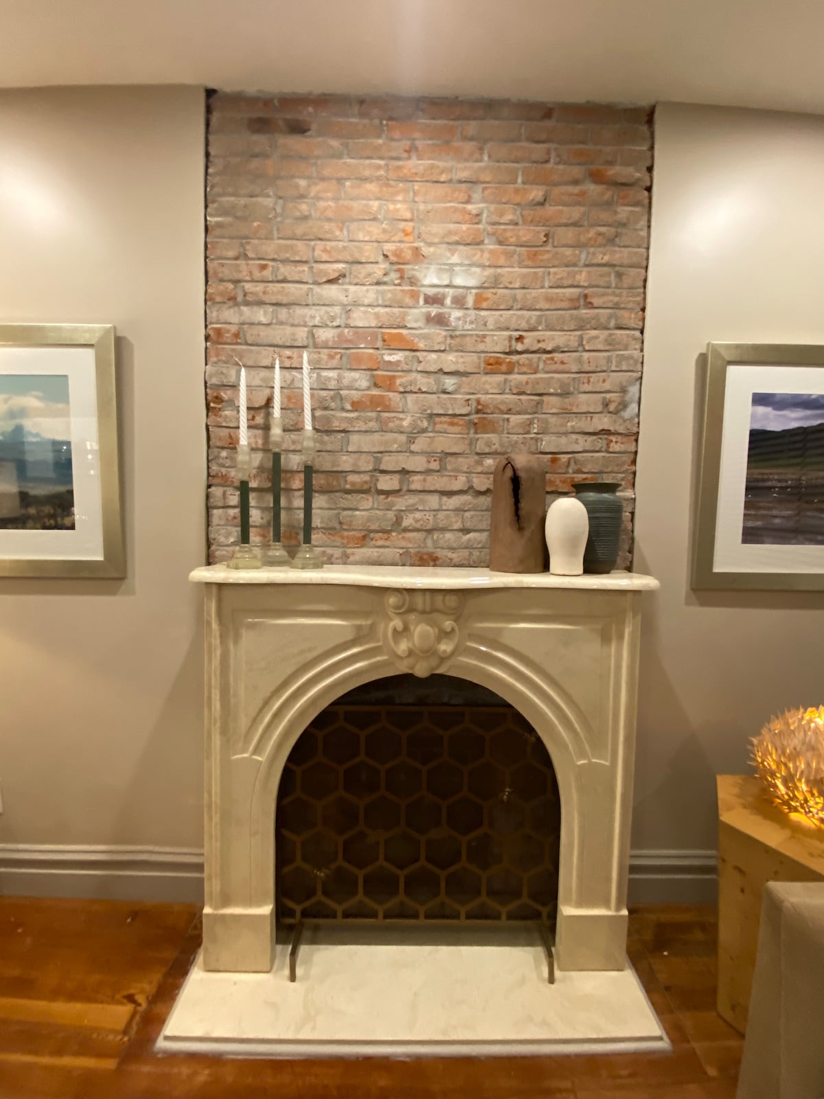 A classic fireplace is framed by exposed brick, contributing to a sophisticated charm. A few decorative candles are positioned on an elegant holder above the mantel. Artwork is displayed on either side, enhancing the space's inviting character.