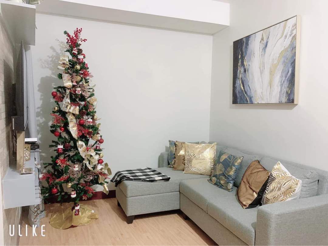 A cozy living space features a light grey sectional sofa adorned with decorative cushions. A Christmas tree, decorated with colorful ornaments and a gold ribbon, stands in the corner, complemented by a wall art piece above. Warm wooden flooring enhances the inviting atmosphere.
