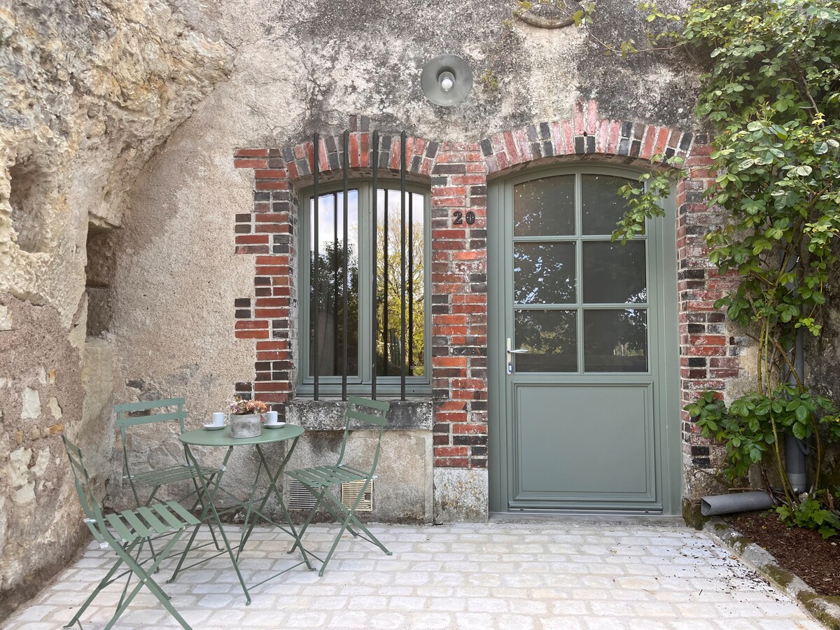 An inviting entrance features a green door framed by rustic brick walls. Two metal chairs and a small table are arranged on a paved patio, surrounded by lush greenery. Large windows offer a glimpse of the outdoors, enhancing the space's charm.
