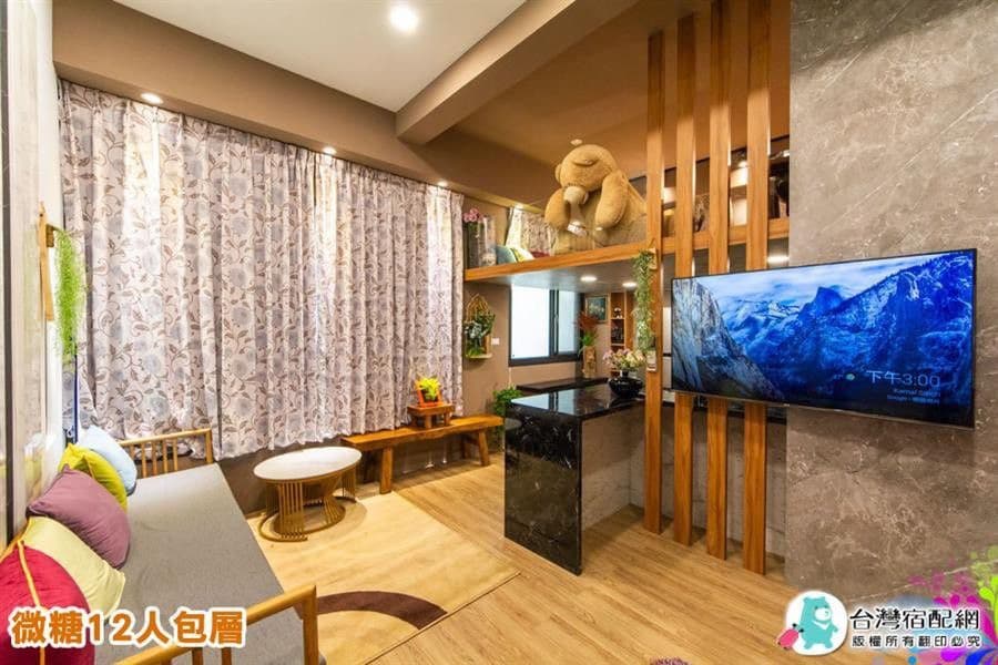 The living area features a cozy seating setup with wooden benches and a round table. A plush toy is positioned on a shelf, while a television mounted on the wall provides entertainment options. Sunlight filters through patterned curtains, accentuating the warm atmosphere.