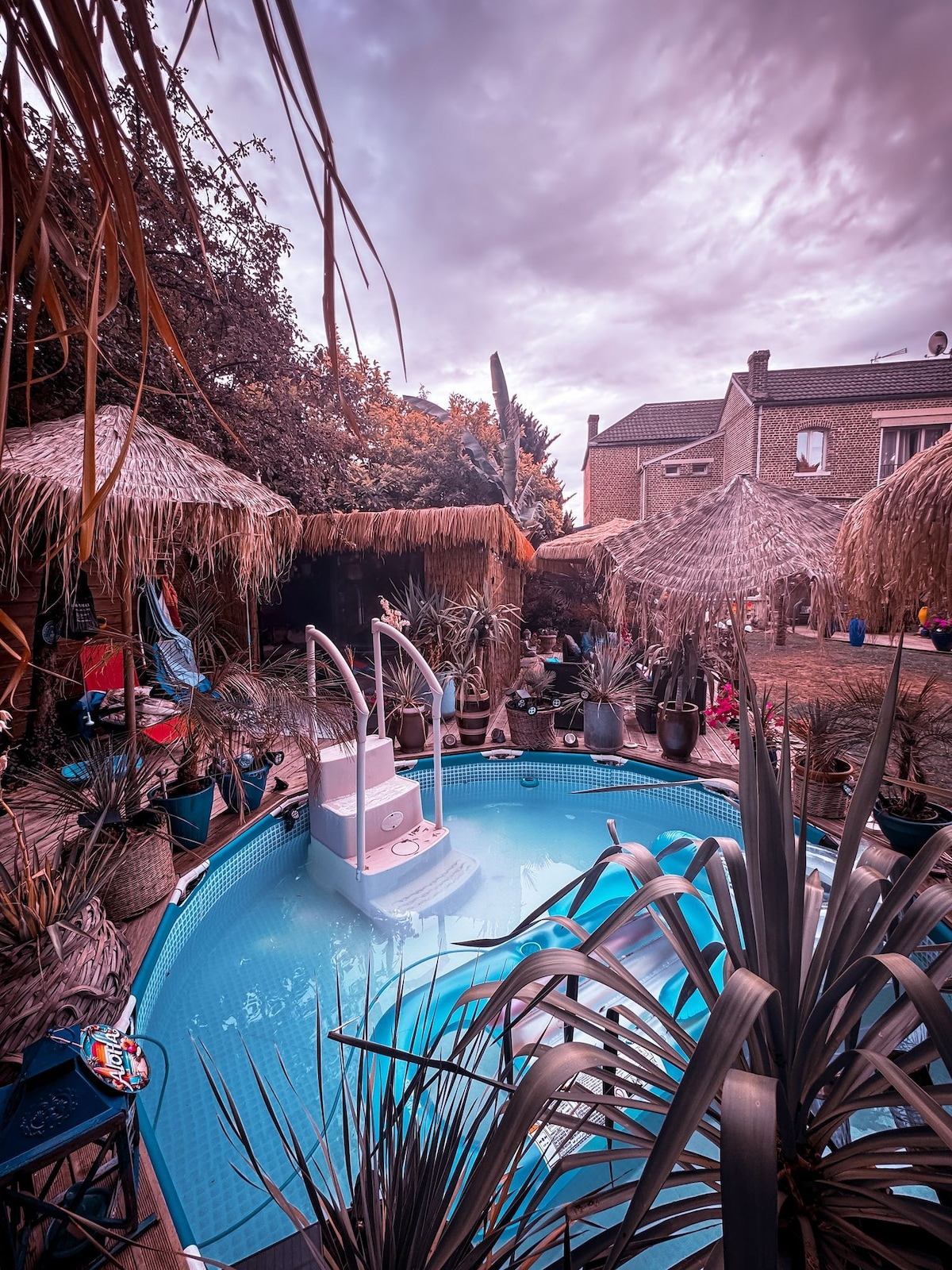 An inviting tub pool is surrounded by thatched umbrellas and lush greenery. A variety of colorful planters enhance the vibrant atmosphere. Lounge chairs are positioned around the pool, creating a relaxed environment. The backdrop features a charming stone building, contributing to the overall serene setting.