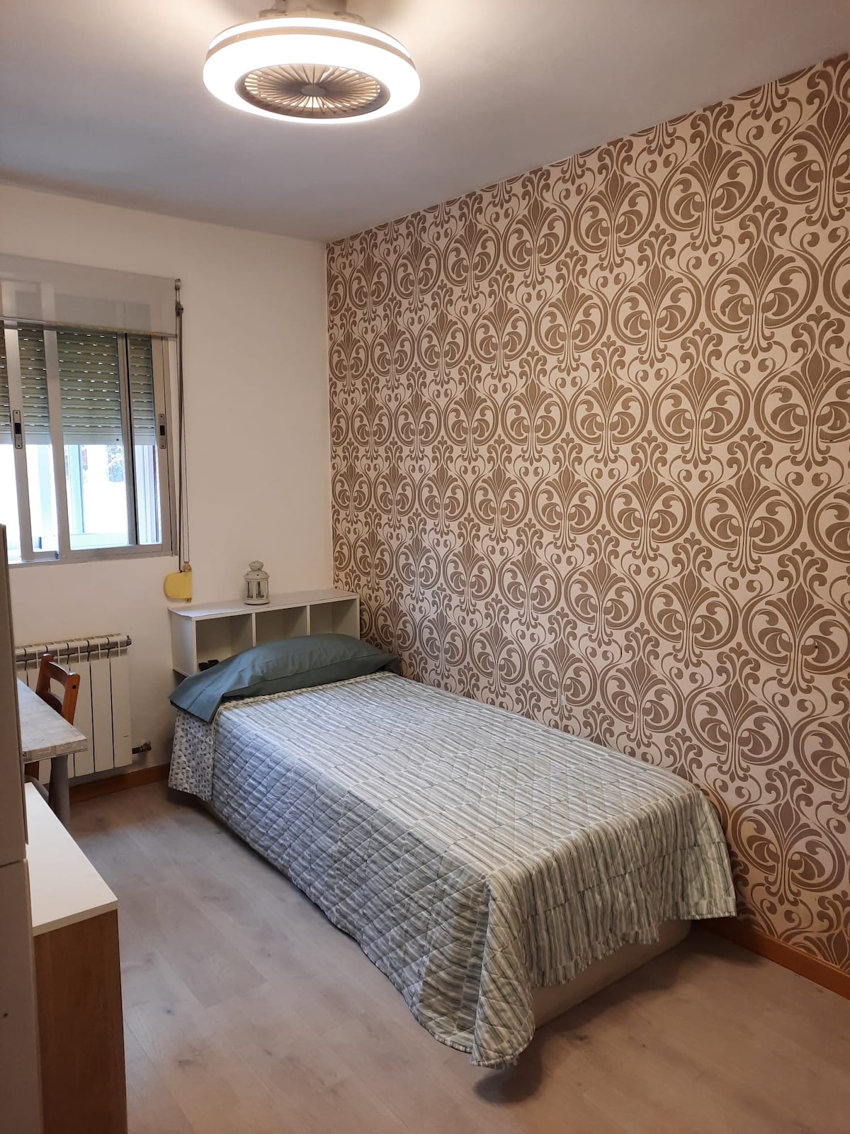 A neatly arranged bedroom is seen, featuring a single bed covered with a light quilt. A patterned wall adds a decorative touch, while a window allows natural light to enter. A small desk and a shelf are included, creating a functional and cozy atmosphere.