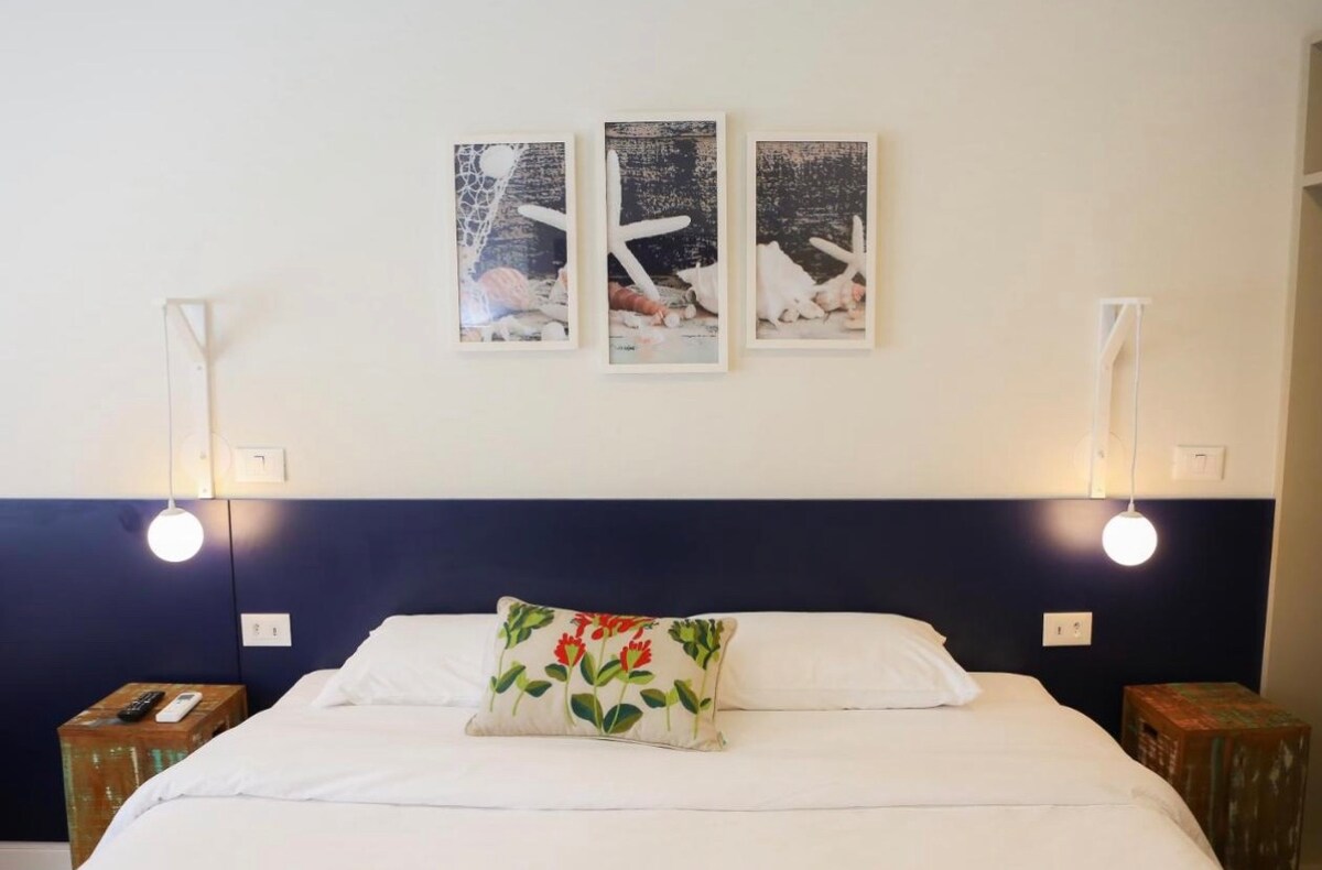 A spacious bed is centered against a navy accent wall, complemented by two decorative side tables. Two round pendant lights provide soft illumination. Three framed artworks hang above the bed, adding visual interest to the serene atmosphere.