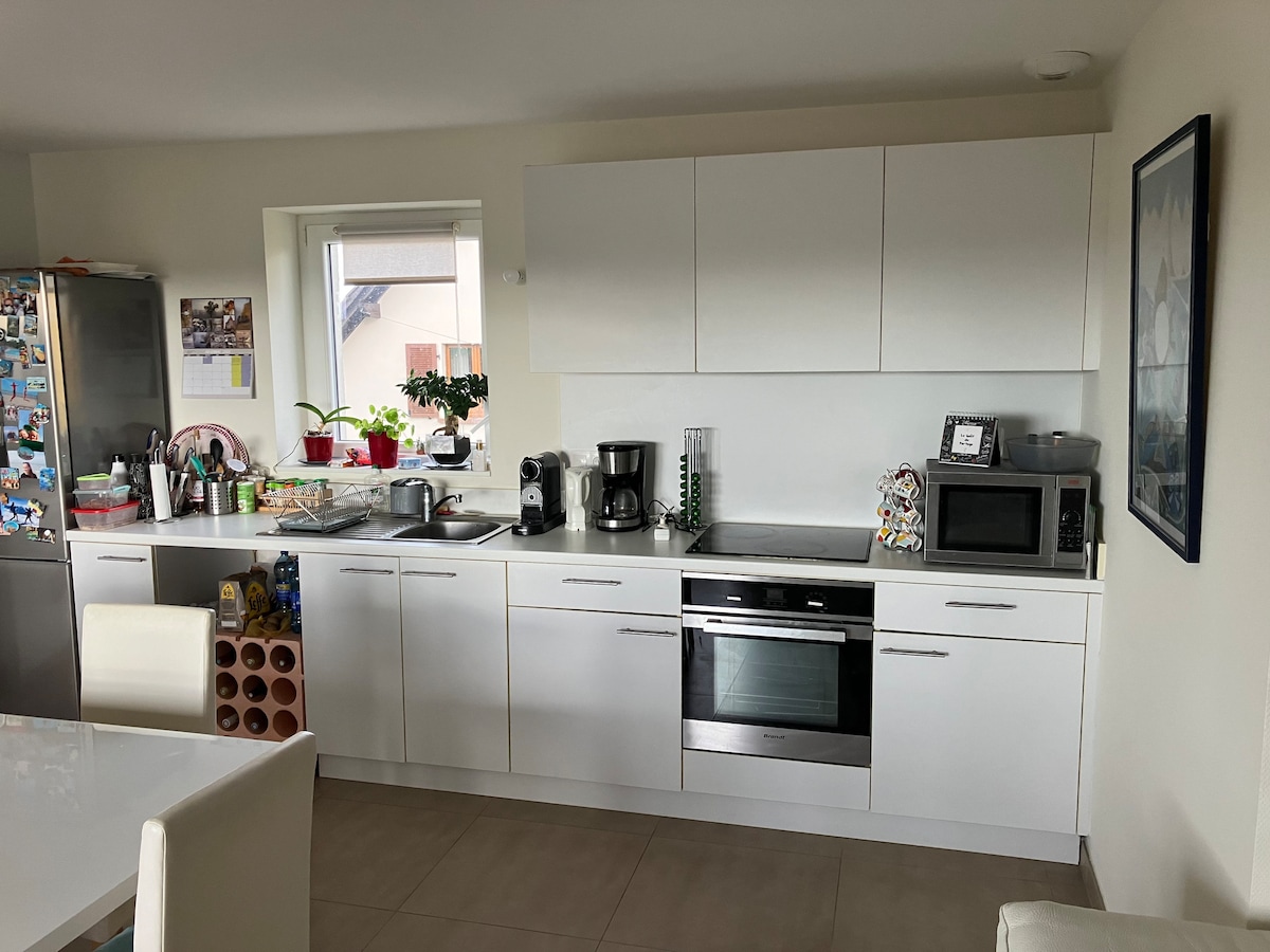 The kitchen features modern white cabinetry and a spacious countertop. Appliances such as a microwave, coffee machine, and oven are included. A window allows natural light to illuminate the area, complemented by potted plants on the sill. A dining table is visible in the foreground.