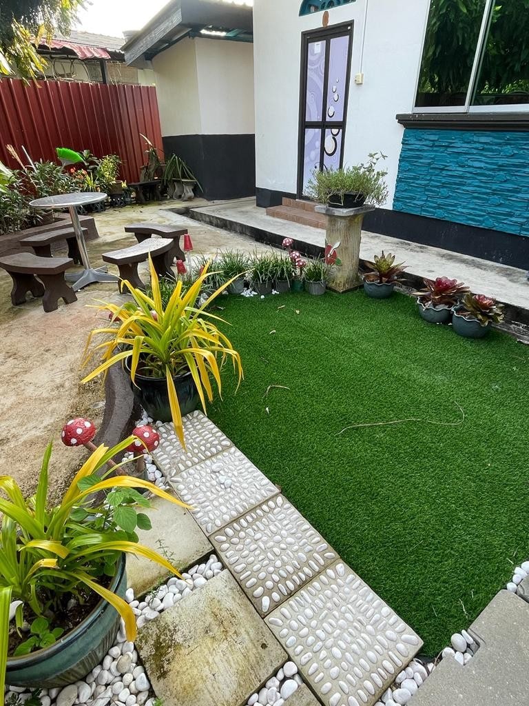 A small outdoor space features artificial grass surrounded by decorative stones and potted plants. Two wooden tables and several chairs create a casual seating area. Greenery and flower arrangements add color to the landscape, while a pathway leads to the entrance of the home.