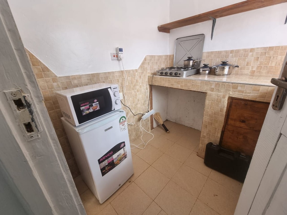 A compact kitchen space features tiled walls and a wooden shelf. Equipped appliances include a microwave and a refrigerator, with a stovetop and several pots on the counter. The floor is tiled, and the area is illuminated by natural light.