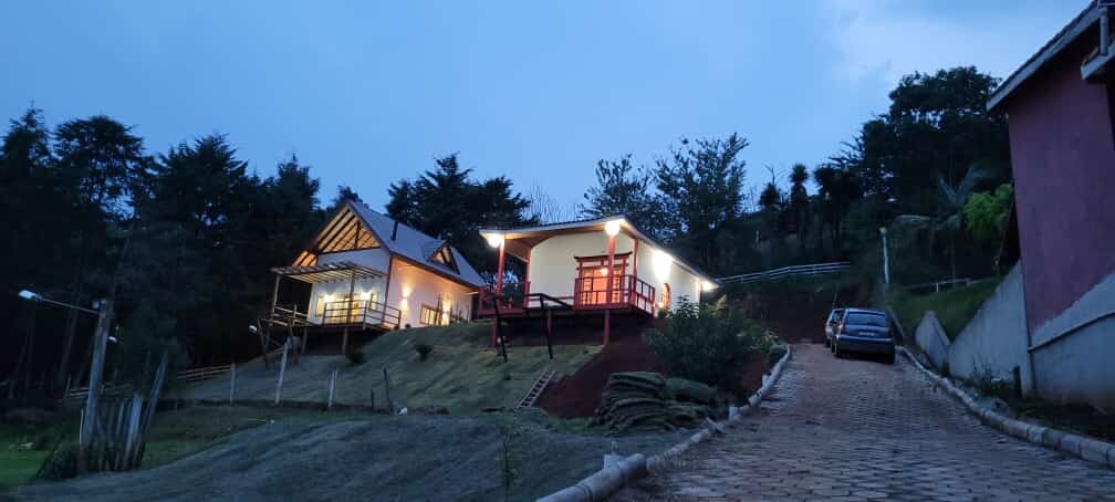 Two accommodation buildings are nestled on a hillside, surrounded by lush greenery. One structure features a traditional Japanese design with a sloped roof, while the other has a modern facade with large windows. Ambient lighting enhances the inviting atmosphere during twilight.