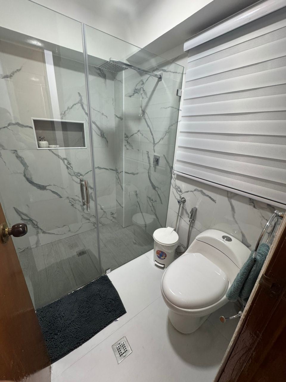 A modern bathroom is displayed, featuring a glass shower enclosure with marble-patterned walls. A toilet is positioned next to a small waste bin. A soft mat lies on the floor, adding comfort and style to the space. Natural light is filtered through a window with blinds.