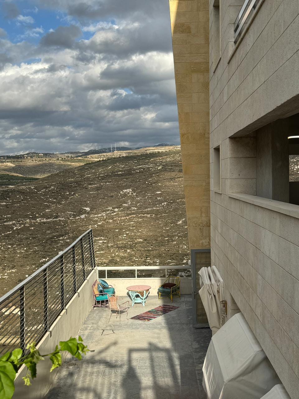 A spacious terrace is visible, adorned with colorful chairs and a small table, overlooking a serene landscape of rolling hills. The stone exterior of the building blends naturally with the environment, while a soft rug adds warmth to the outdoor space.