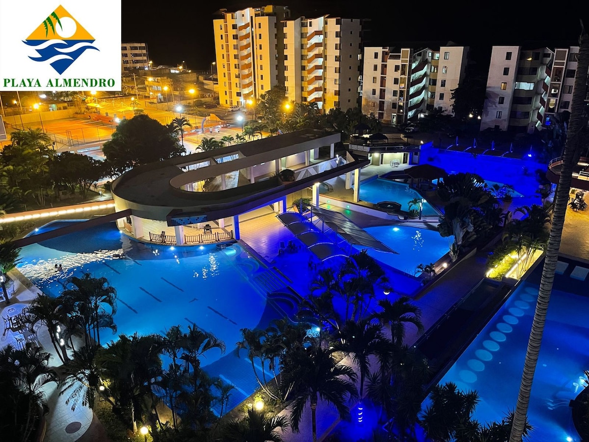 A large outdoor pool area is illuminated in blue light, surrounded by lush palm trees. Several lounge chairs are visible along the edge of the pools. Multiple white buildings can be seen in the background, contributing to the setting's family-friendly atmosphere.