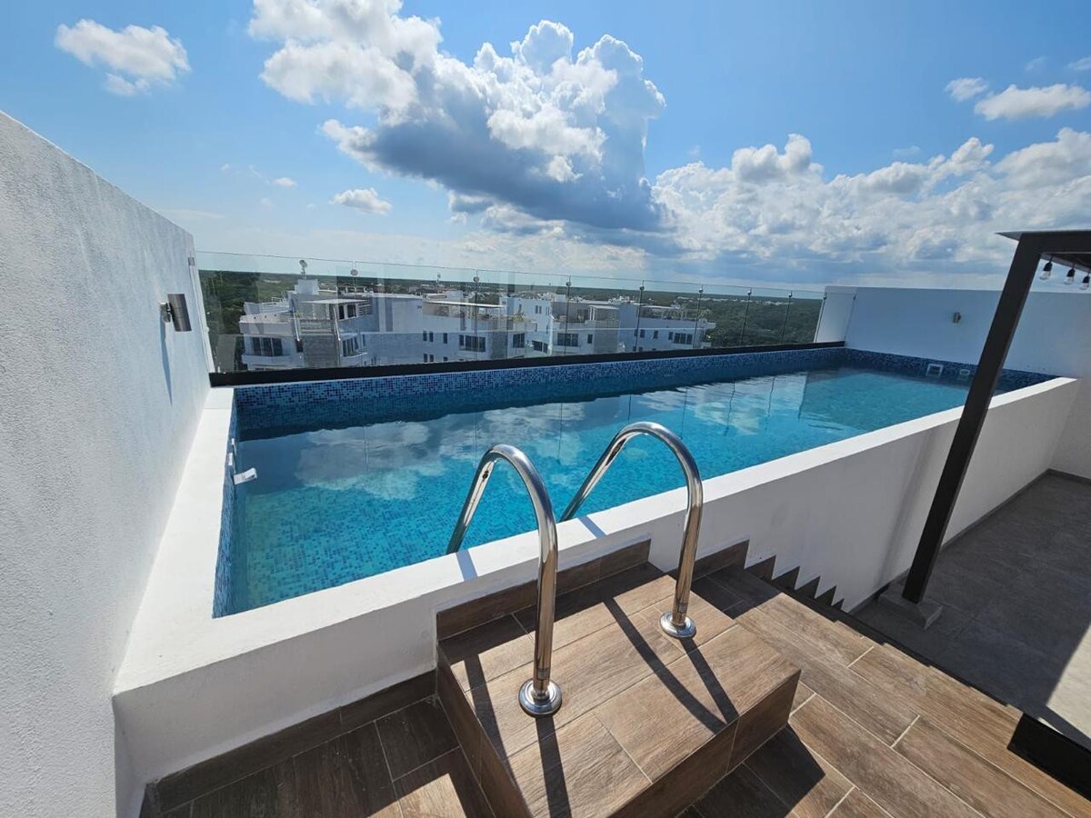 An inviting rooftop pool features clean lines and a stunning view of the surrounding area. The poolside is equipped with a set of stainless steel steps, and the serene water is framed by a glass railing, enhancing the open atmosphere.