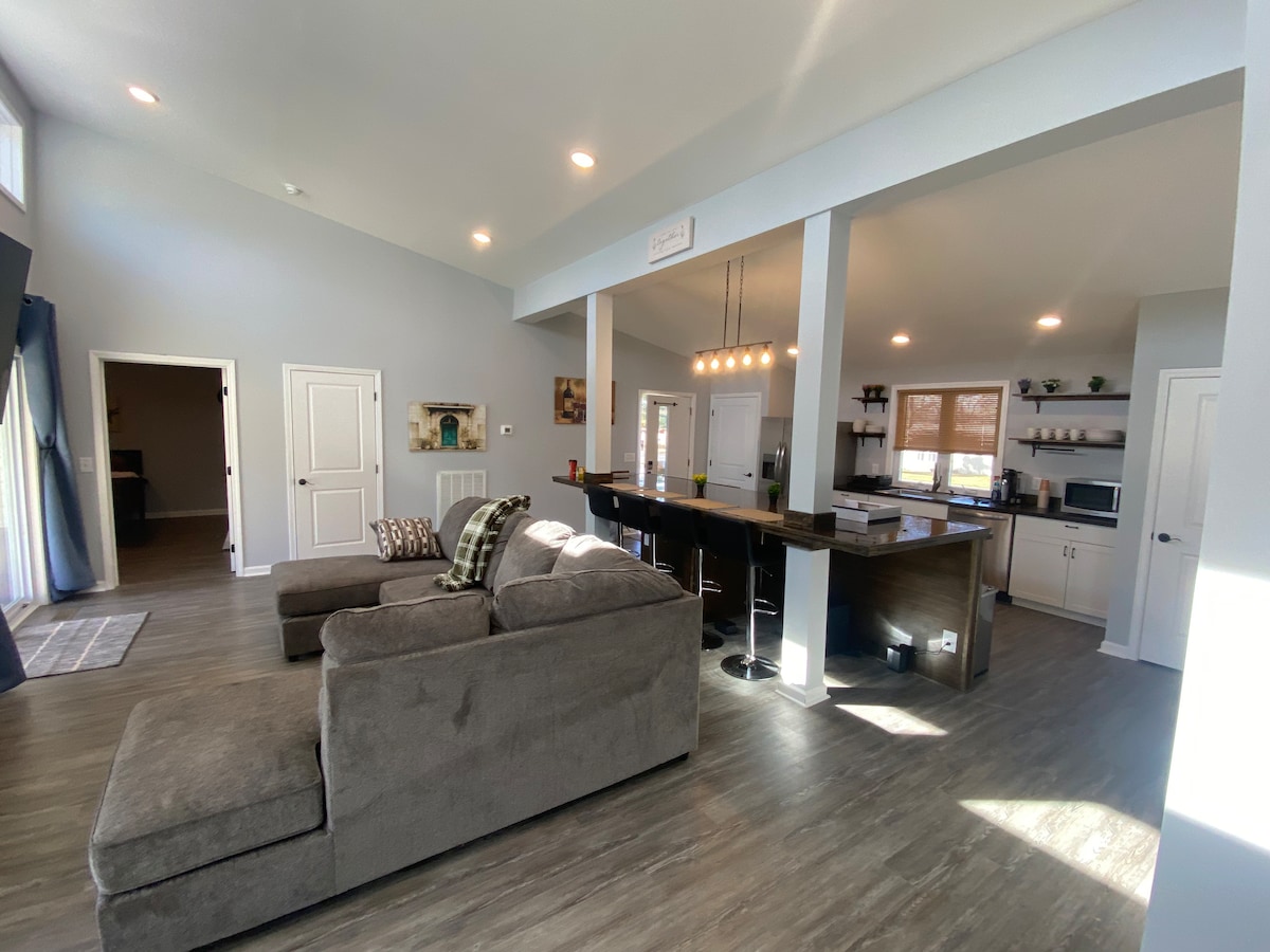 An open living area is illuminated by natural light, featuring a large, comfortable sectional sofa. The adjacent kitchen boasts a bar with seating and modern appliances. The walls are painted in soft tones, enhancing the spacious feel, while doors lead to other parts of the home.