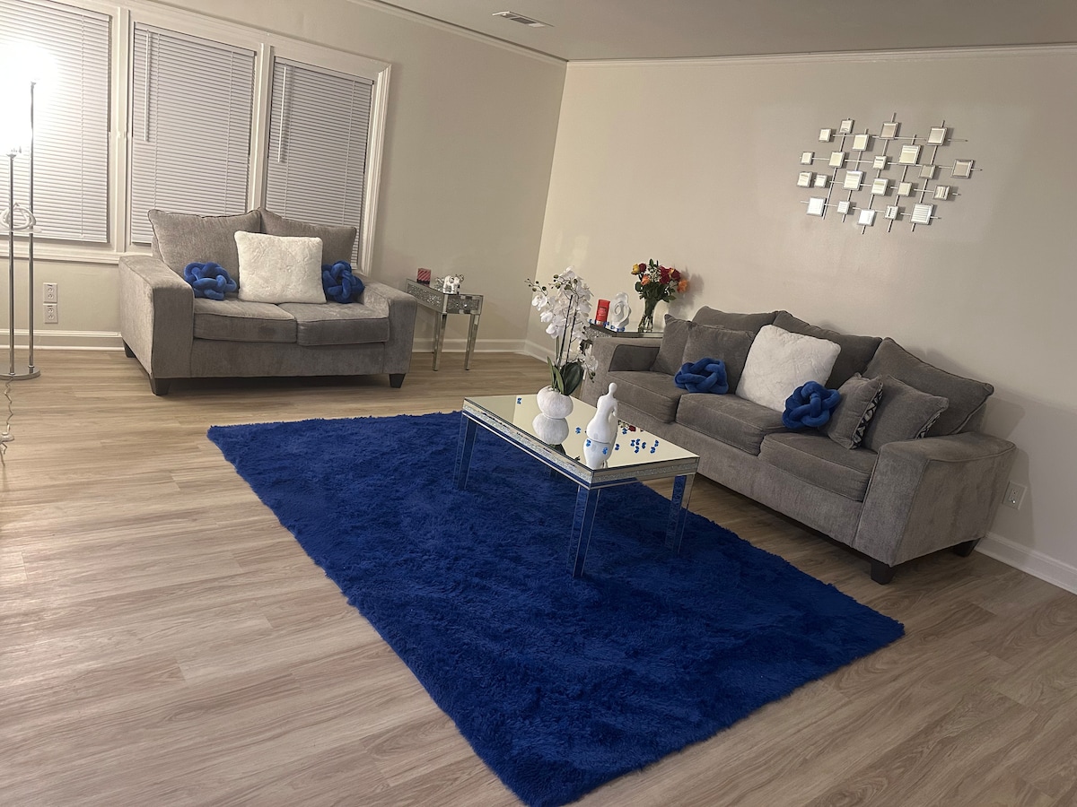 The living room features two gray sofas adorned with decorative pillows, positioned around a glass coffee table. A plush blue rug adds warmth to the space, while a cluster of decorative items sits atop the table. Large windows allow natural light to fill the area.